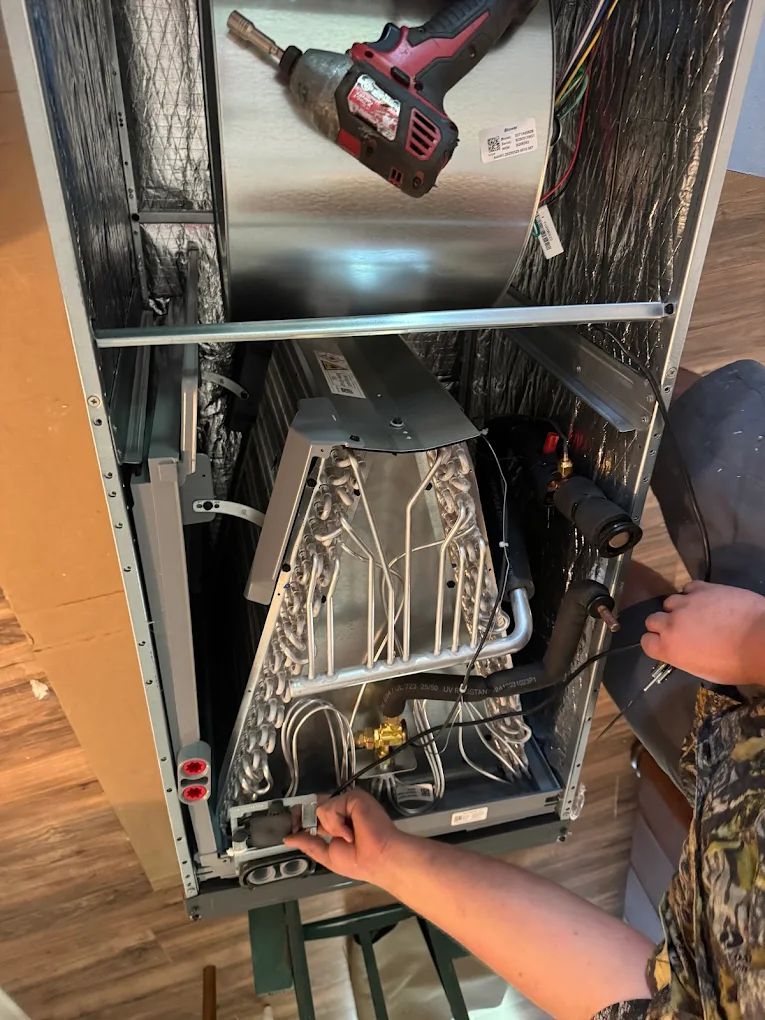 Person working on an HVAC unit. Inside a rectangular frame are components, including a coil. A power drill is overhead.