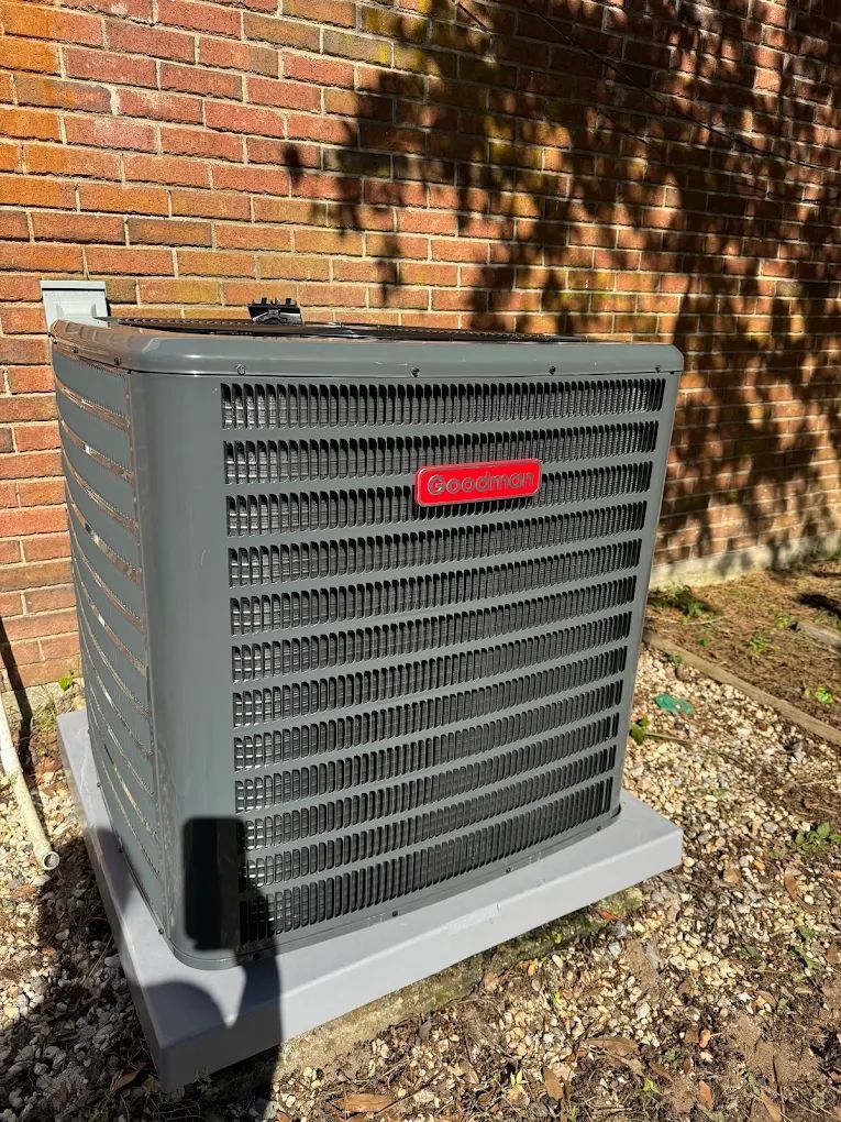 Gray Goodman air conditioner unit against a brick wall, sitting on a concrete pad in the yard.
