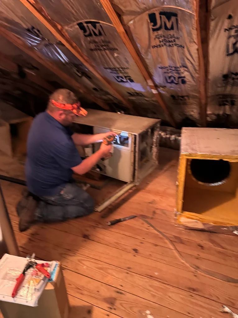 Man working on HVAC ductwork in an attic. He's kneeling, wearing a headlamp, surrounded by ducts and insulation.
