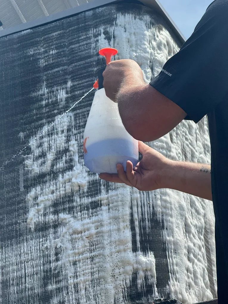Person spraying a foamy cleaner on a black air conditioning unit outdoors.