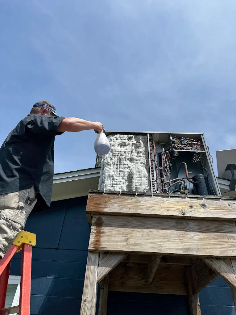 Man on ladder spraying the inside of an air conditioning unit on a rooftop with a blue sky overhead.