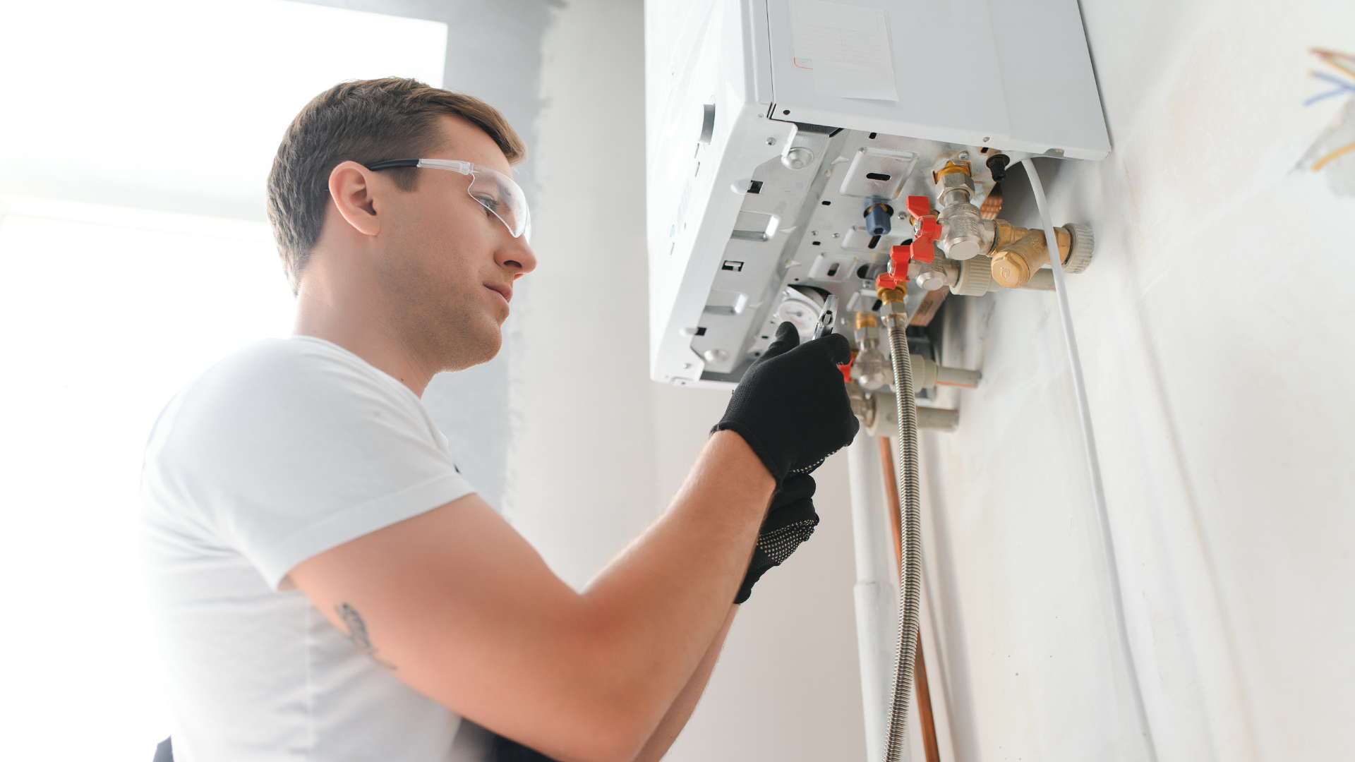 A man in safety glasses and gloves repairs plumbing on a white wall.