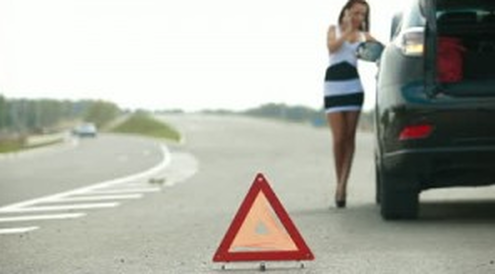 A woman is standing next to a broken down car and talking on a cell phone.