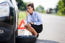 A woman is squatting next to a tire and talking on a cell phone.