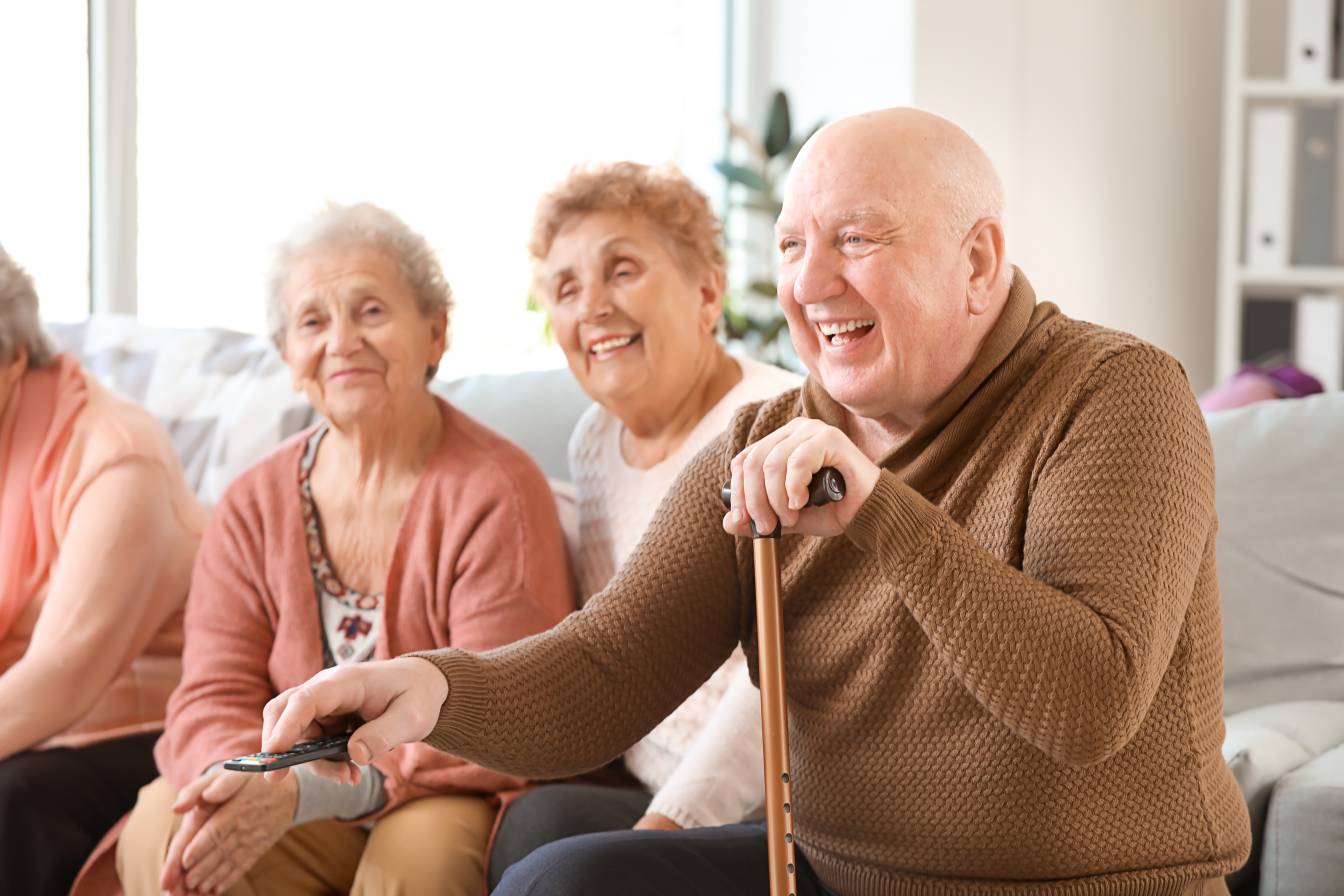 An old man and two old women laughing at something on TV in a care home