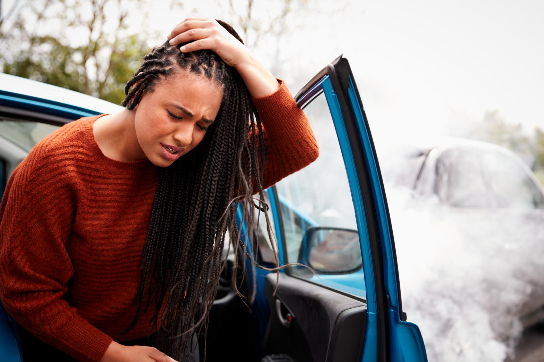 Woman with braided hair injured after car accident; she is holding her head, near her damaged car.