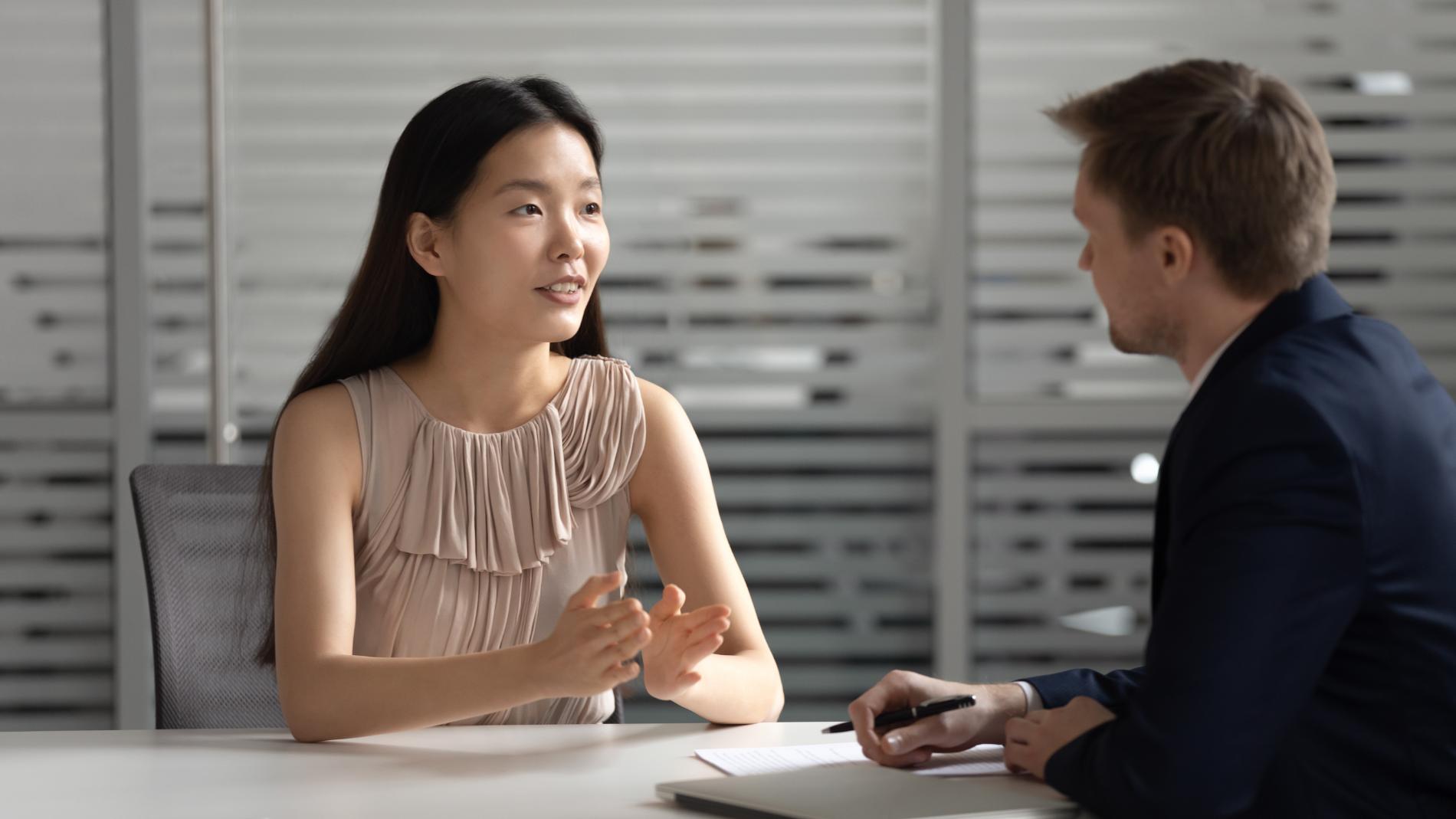 Woman in beige top gestures while talking to a man in a suit at a desk in an office.
