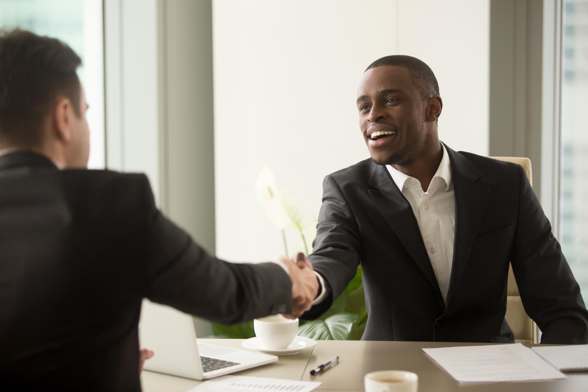 Two men shaking hands in an office setting. Black man smiling, wearing a suit, sitting at a desk.