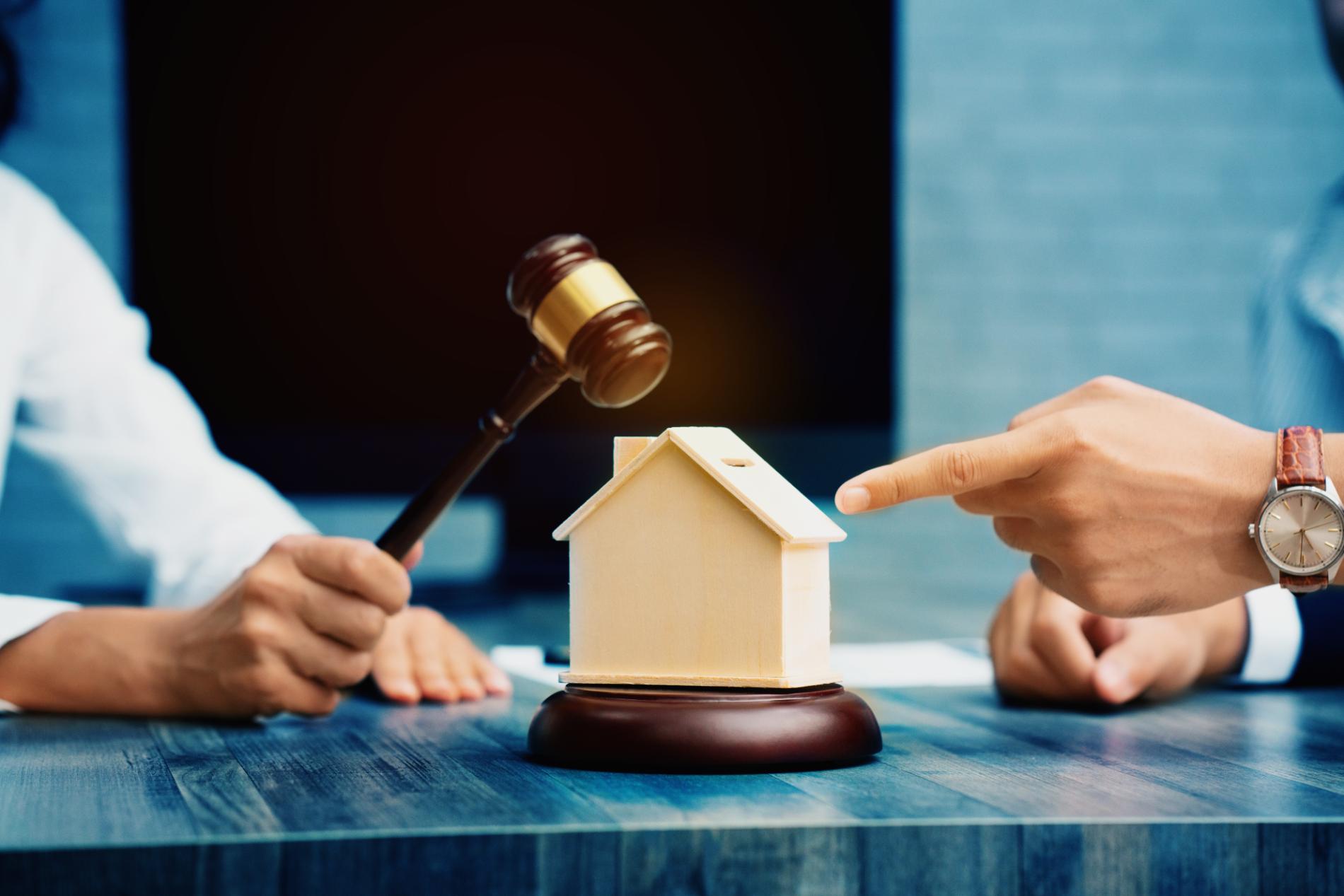 Hands holding a gavel and pointing to a small house model on a table, suggesting a property auction.