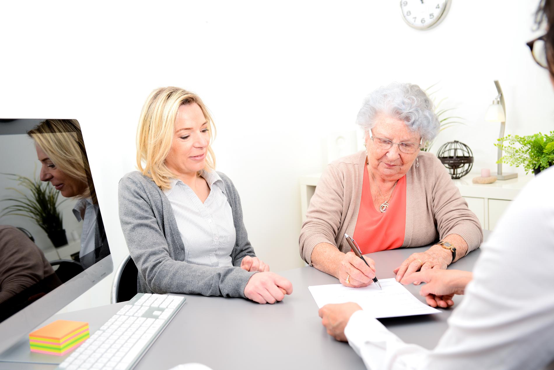 Woman signing document with another woman and a person holding the document, in an office setting.