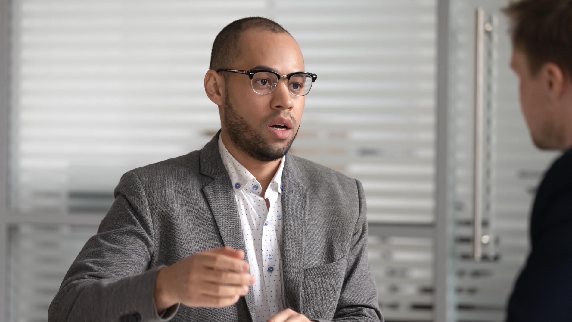 Man in glasses gestures while speaking to another man in an office.