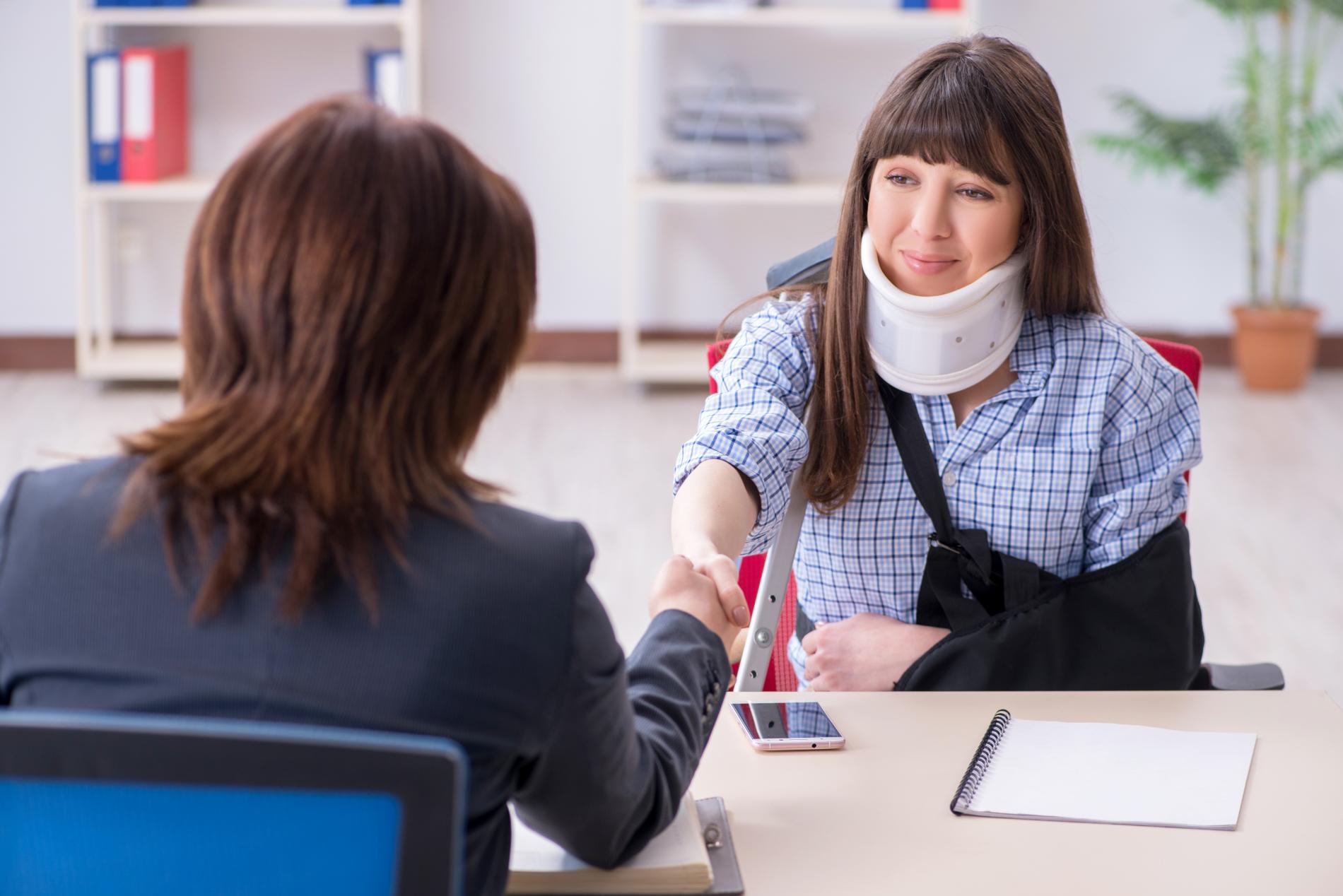 Woman with neck brace and arm sling shaking hands with a woman in an office setting.