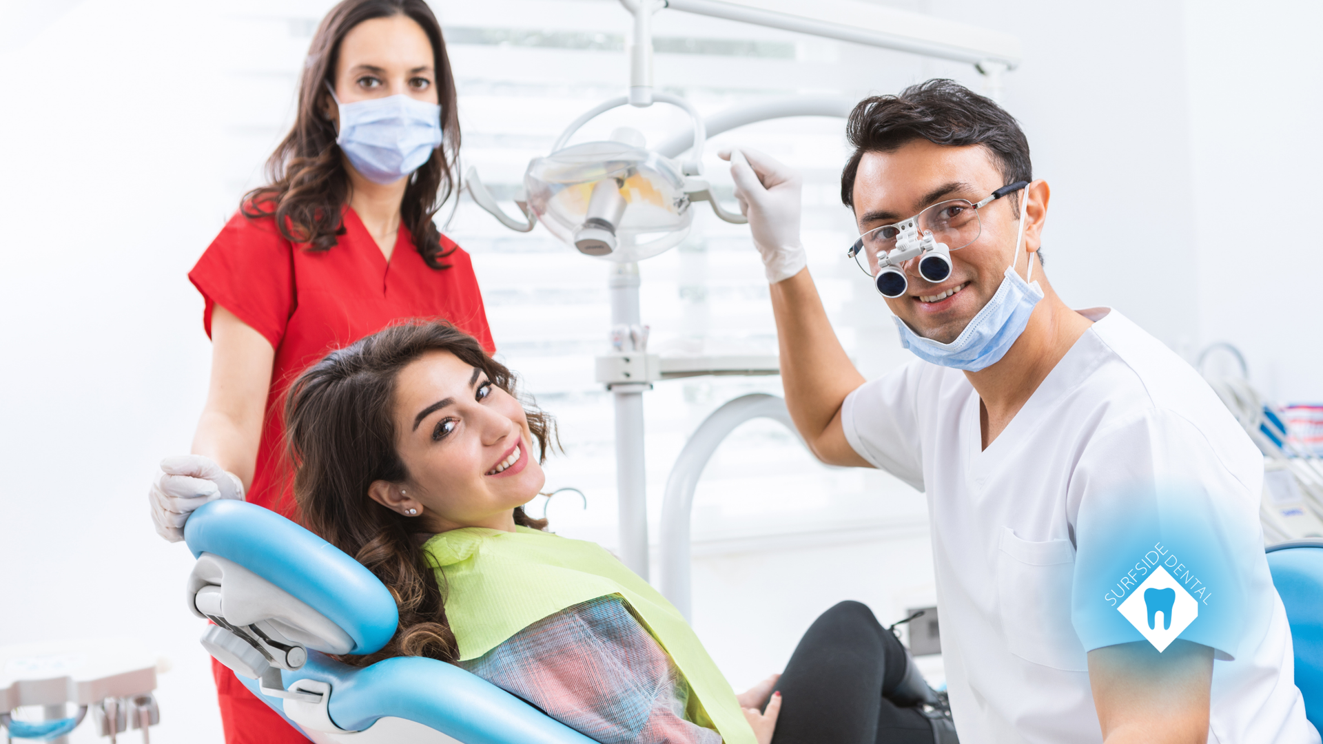 Dentist, assistant, and patient smiling in a dental office. Patient in chair, dentist wearing glasses, assistant in red.