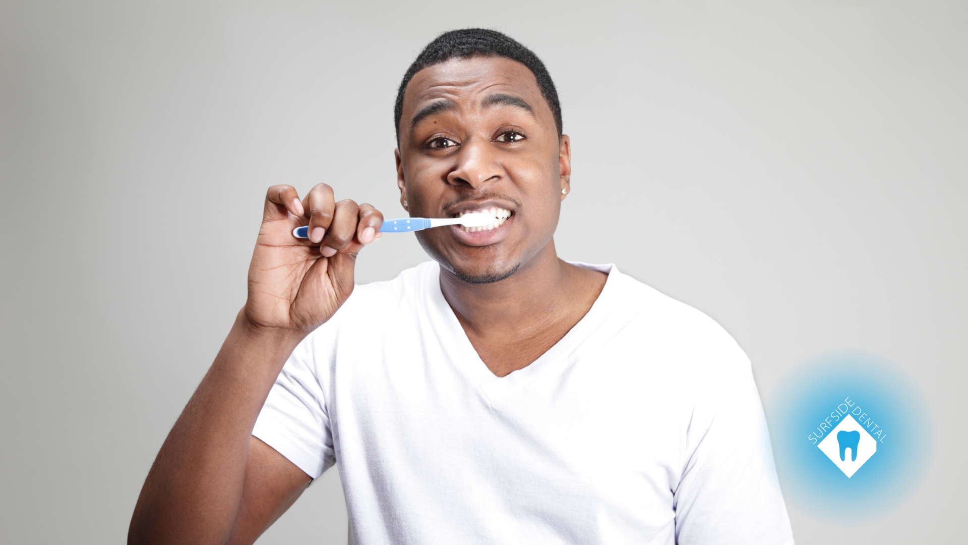 Man brushing his teeth, wearing a white shirt, against a gray backdrop.