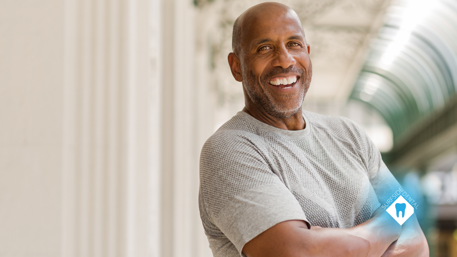 Smiling Black man with crossed arms, wearing a gray shirt, outside.