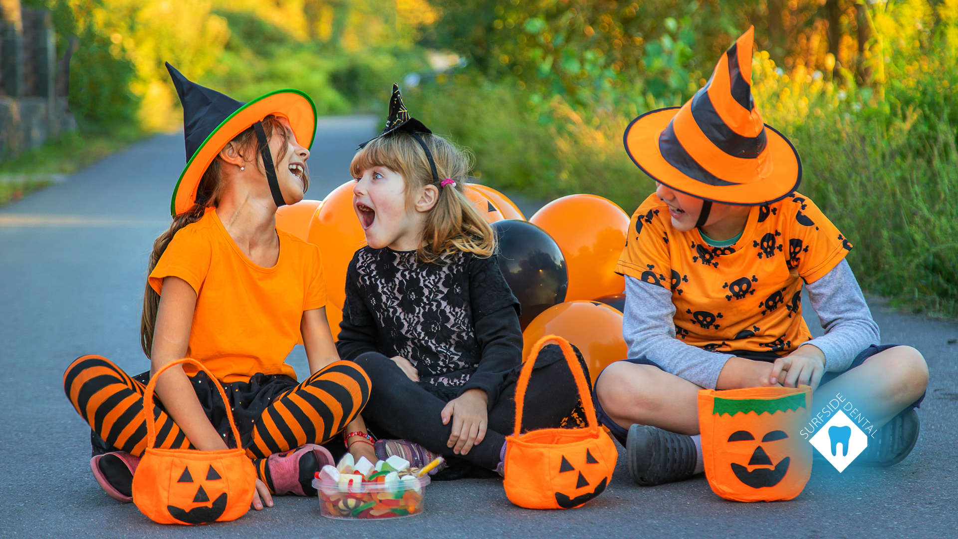 Three children in Halloween costumes sit on a road with jack-o'-lantern candy buckets, orange balloons, and candy.