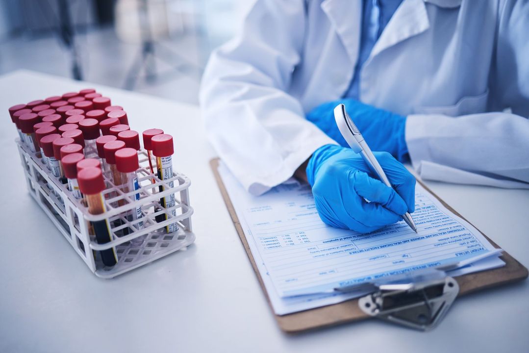 Lab technician in blue gloves, writing on a clipboard with blood sample tubes in a rack on a white lab table.