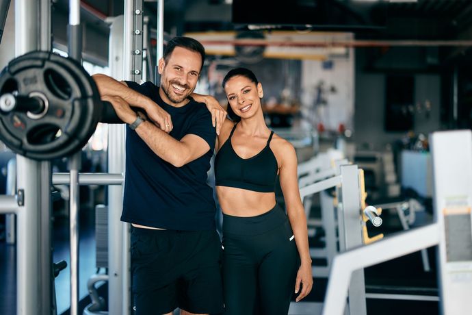 Man and woman smiling at a gym; man leans on barbell, woman rests arm on man.