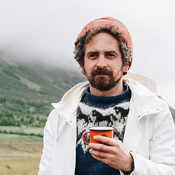 Man with beard in hat and sweater holding orange mug in mountainous setting.