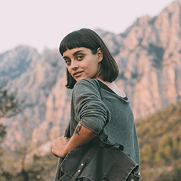 Woman with dark bob, arm tattoo, looking back over shoulder. Mountain backdrop.