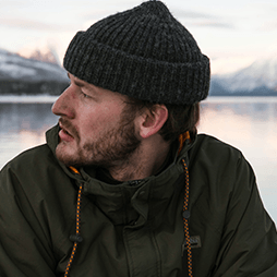 Man in beanie and green jacket looking off-camera; snowy mountain lake background.