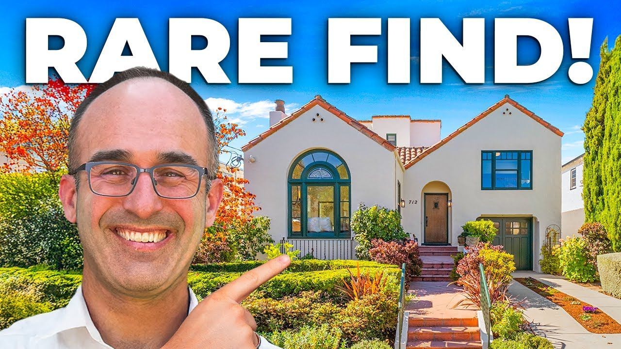 Man pointing at a Spanish-style house; sunny day with blue sky, green plants, and a driveway.