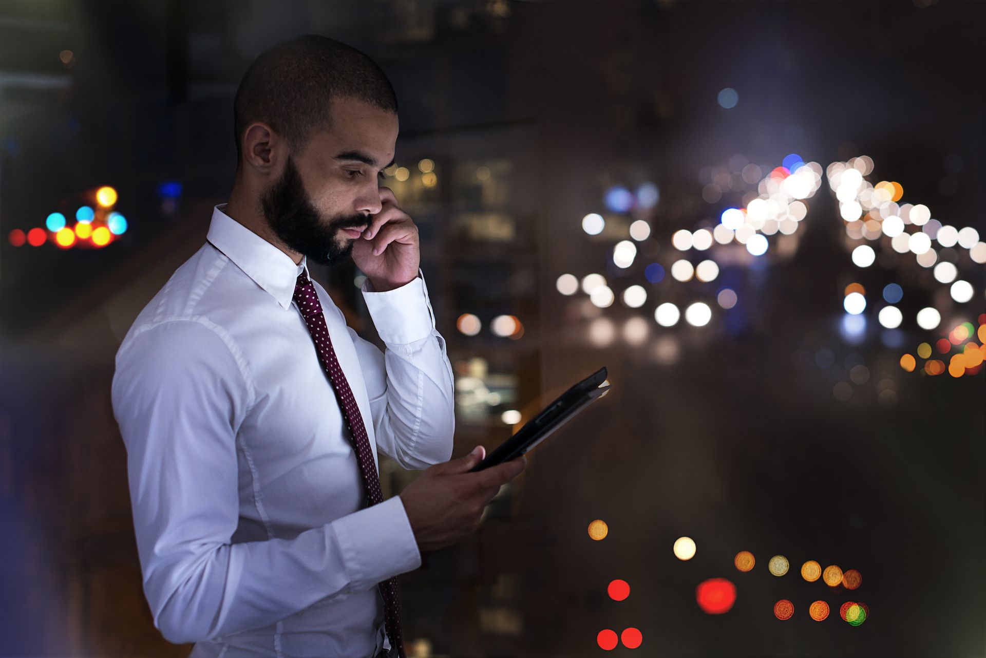 A man with a beard is looking at a tablet while talking on a cell phone.