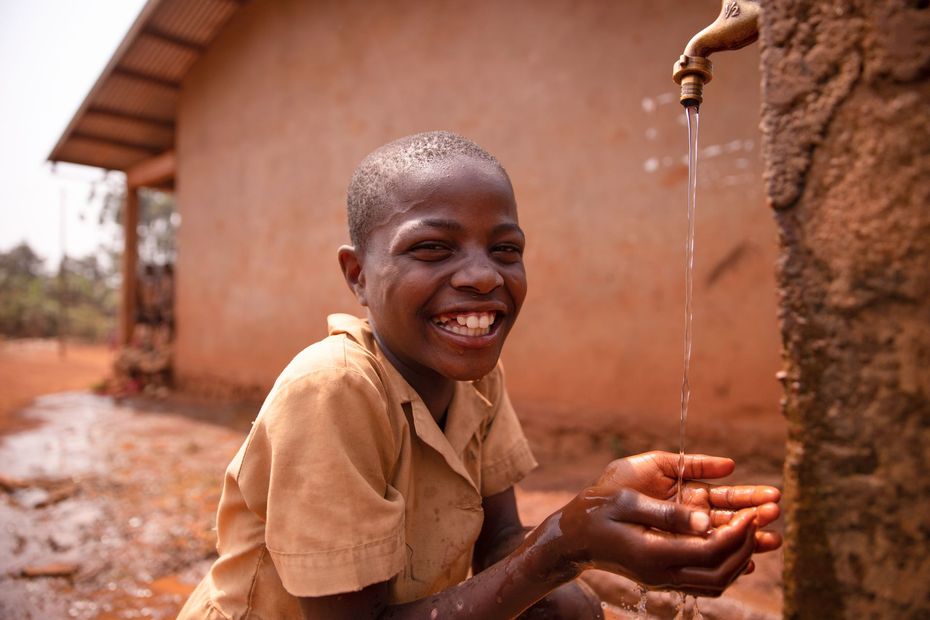 A young boy is washing his hands under a faucet and smiling.