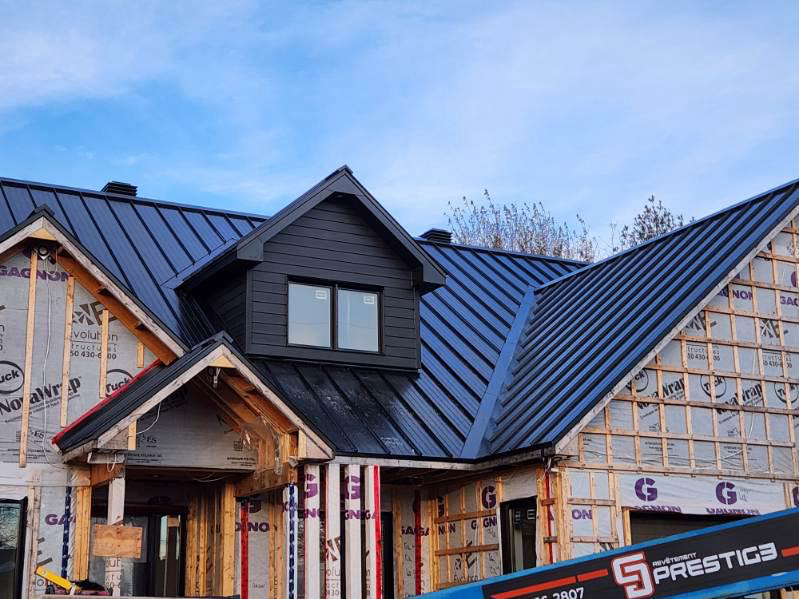 Maison en construction avec toit en métal noir, lucarne et charpente apparente en bois.