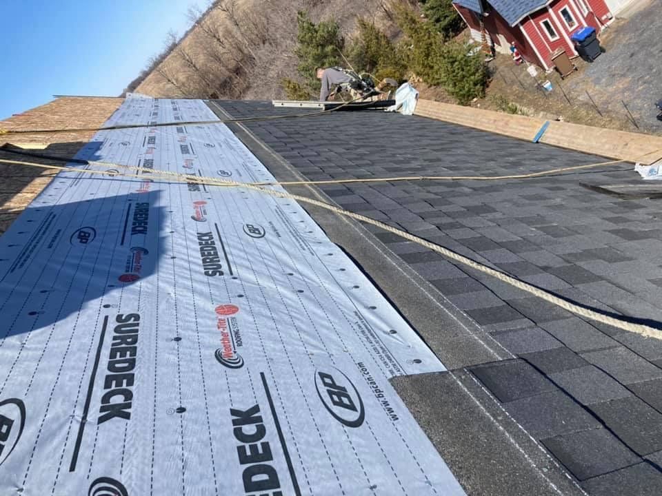 Un toit avec des bardeaux en cours d'installation et une maison rouge en arrière-plan.