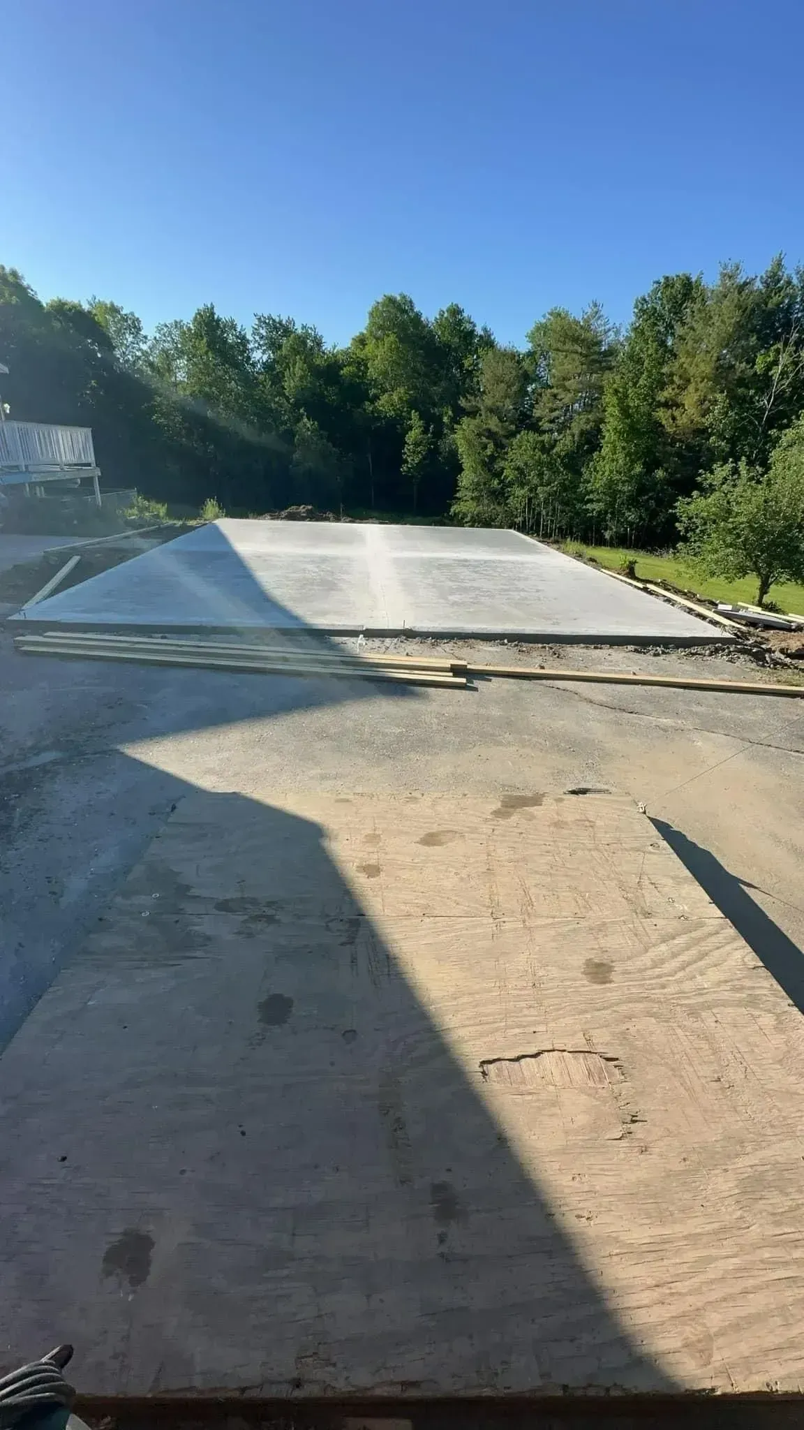 A newly poured concrete pad in a gravel area, with trees in the background under a blue sky.