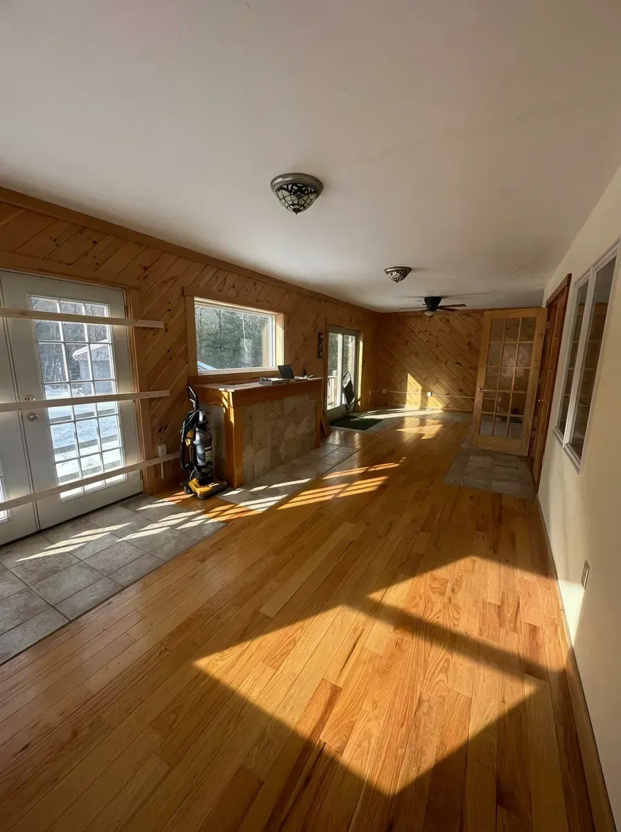 A bright room with wood-paneled walls, hardwood floors, a stone fireplace, and French doors leading outdoors.