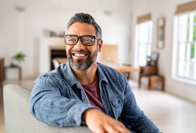 A man is sitting on a couch wearing glasses and smiling.