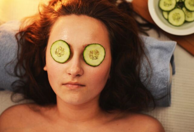 A woman is laying on a bed with cucumber slices on her eyes.