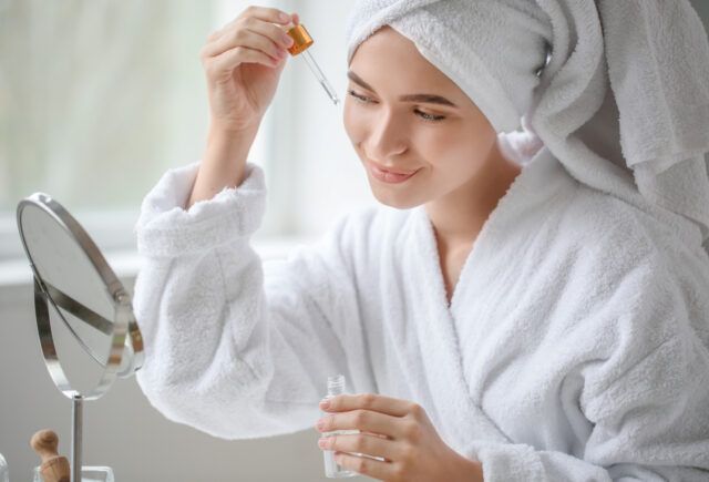 A woman with a towel wrapped around her head is applying a serum to her face in front of a mirror.