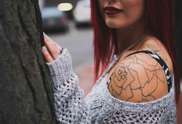 A woman with red hair has a tattoo of a rose on her shoulder.