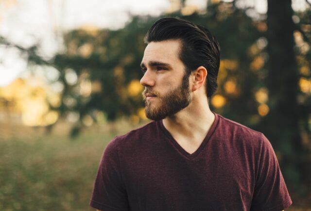 A man with a beard is wearing a maroon t-shirt and standing in the woods.