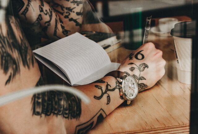 A man with tattoos on his arm is writing in a notebook.