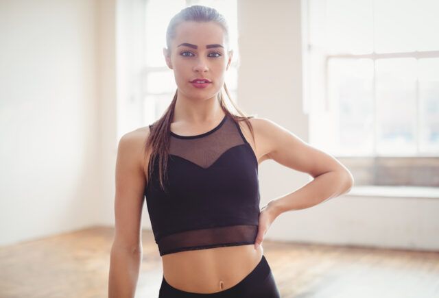 A woman in a black crop top and black leggings is standing in a room with her hands on her hips.