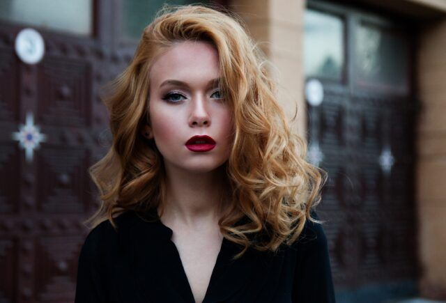 A woman with red hair and red lipstick is standing in front of a building.