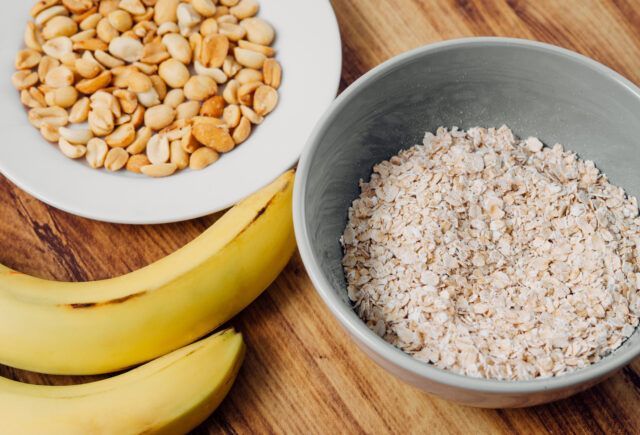 A bowl of oatmeal next to a bowl of peanuts and bananas on a wooden table.