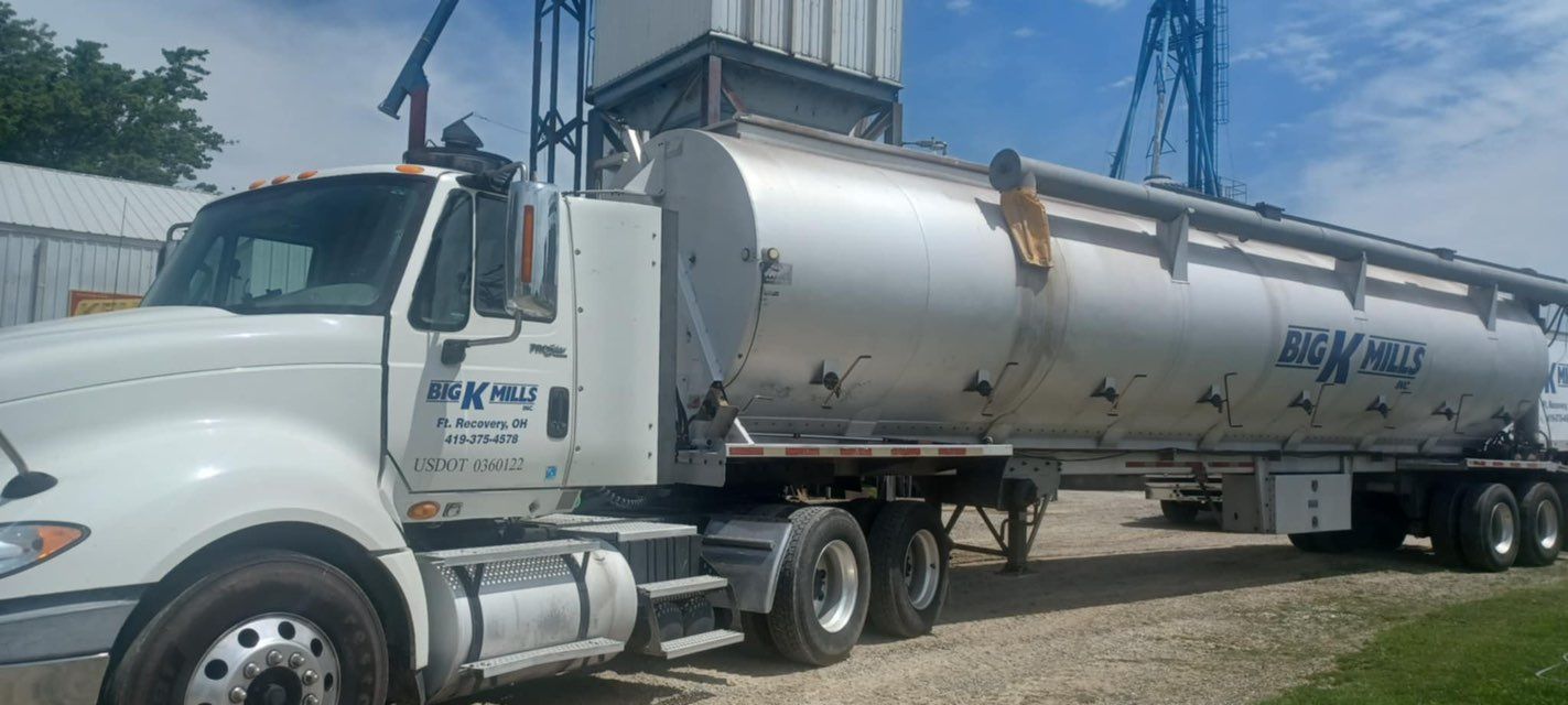 White tanker truck parked near a metal structure on a gravel lot. The truck has the logo