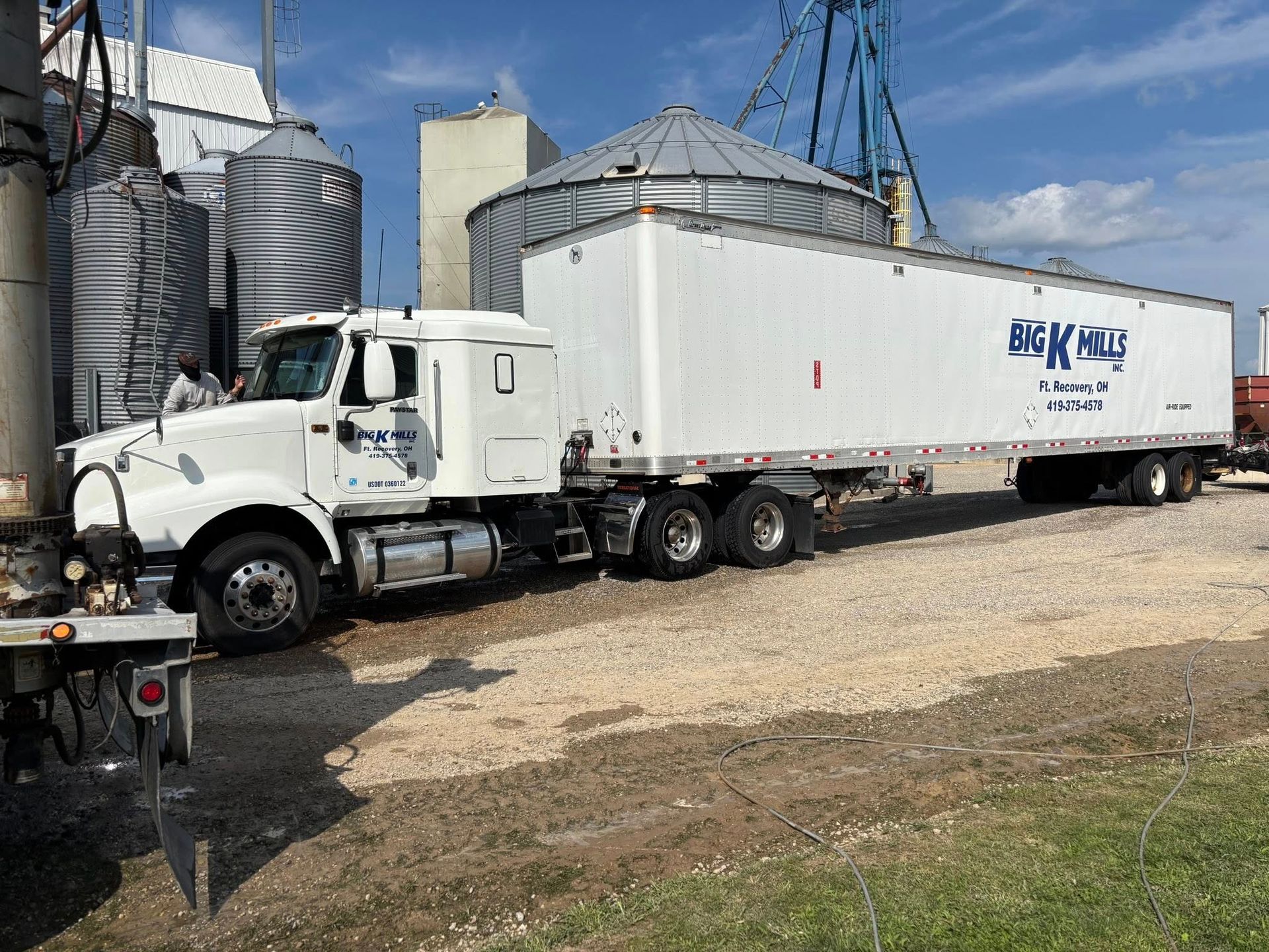 White semi-truck with trailer at grain silo. Someone is standing near the truck. Gravel ground, blue sky.