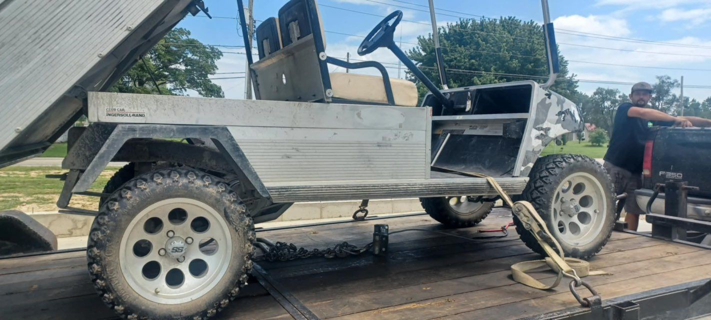 A modified golf cart with large tires is being loaded onto a flatbed truck. A person is assisting.