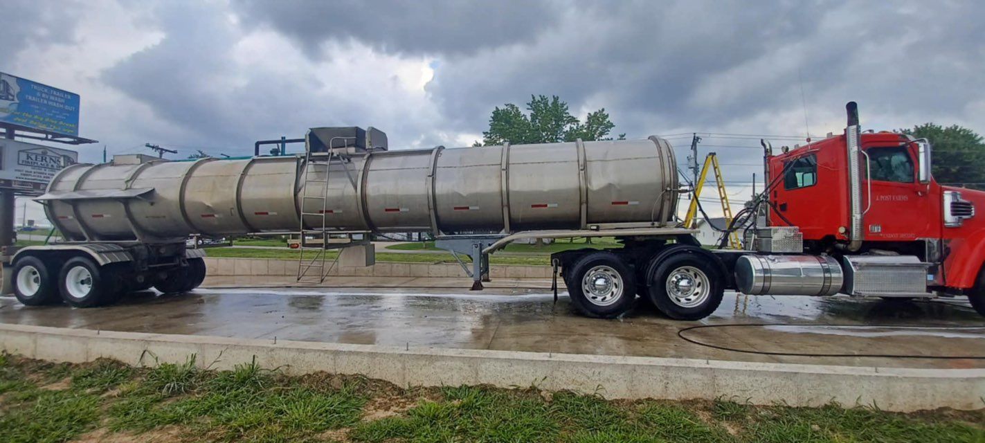 Red semi-truck with a silver tank trailer parked on a wet surface under an overcast sky.