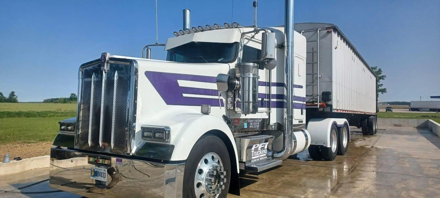 White semi-truck with purple stripes and chrome grill on a concrete surface with field and blue sky in the background.