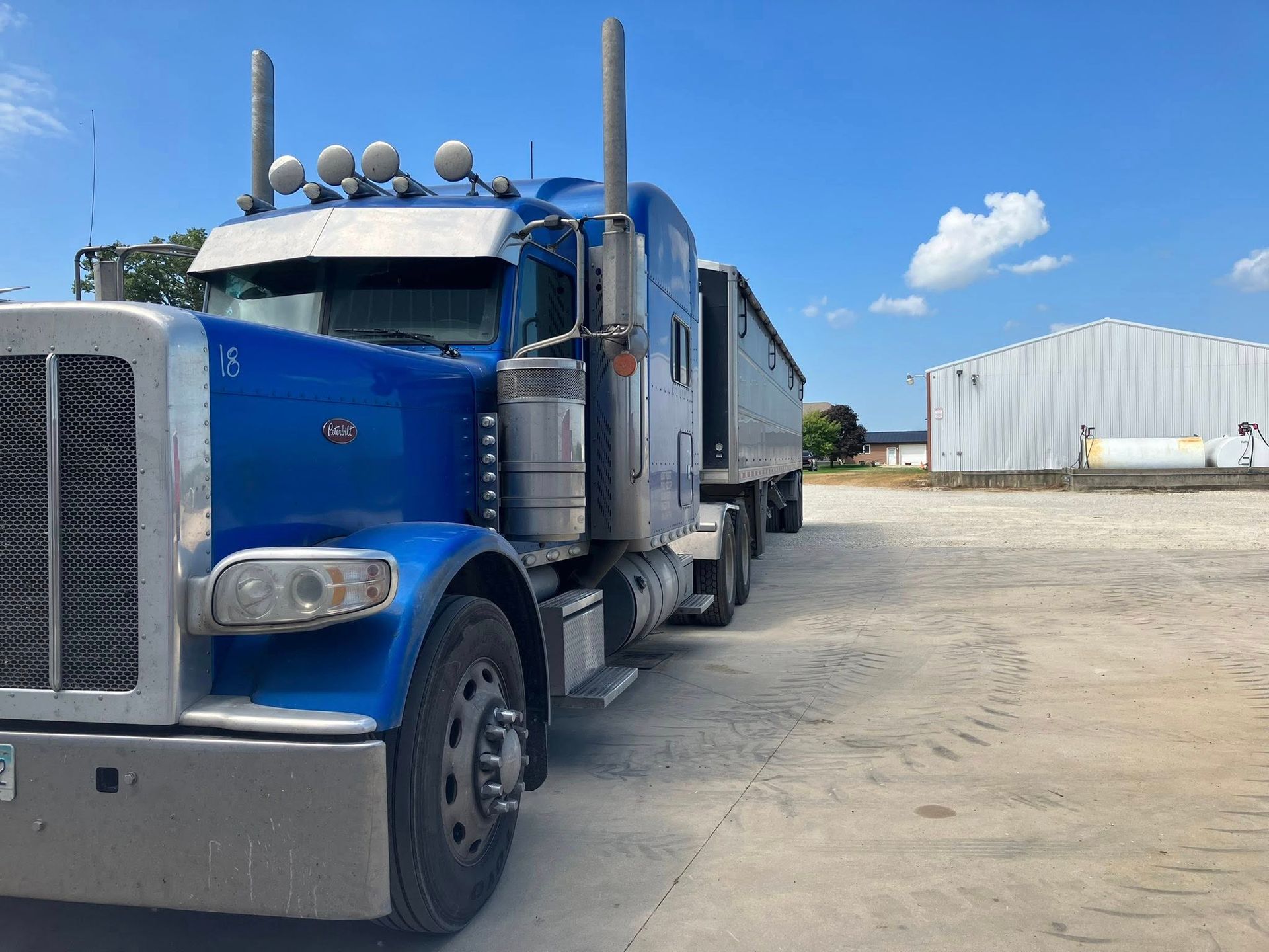 Blue semi-truck with a trailer parked in a lot under a blue sky, next to a white building.