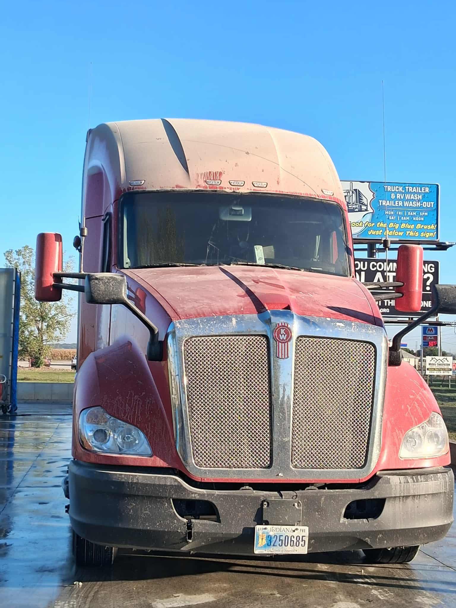 Red semi-truck with a dirty exterior parked in front of a blue sky and various signs.