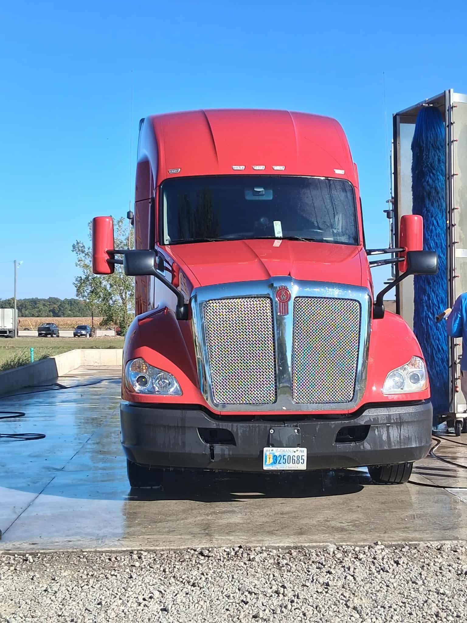 Red semi-truck in a truck wash, the grill is silver, blue cleaning brushes.