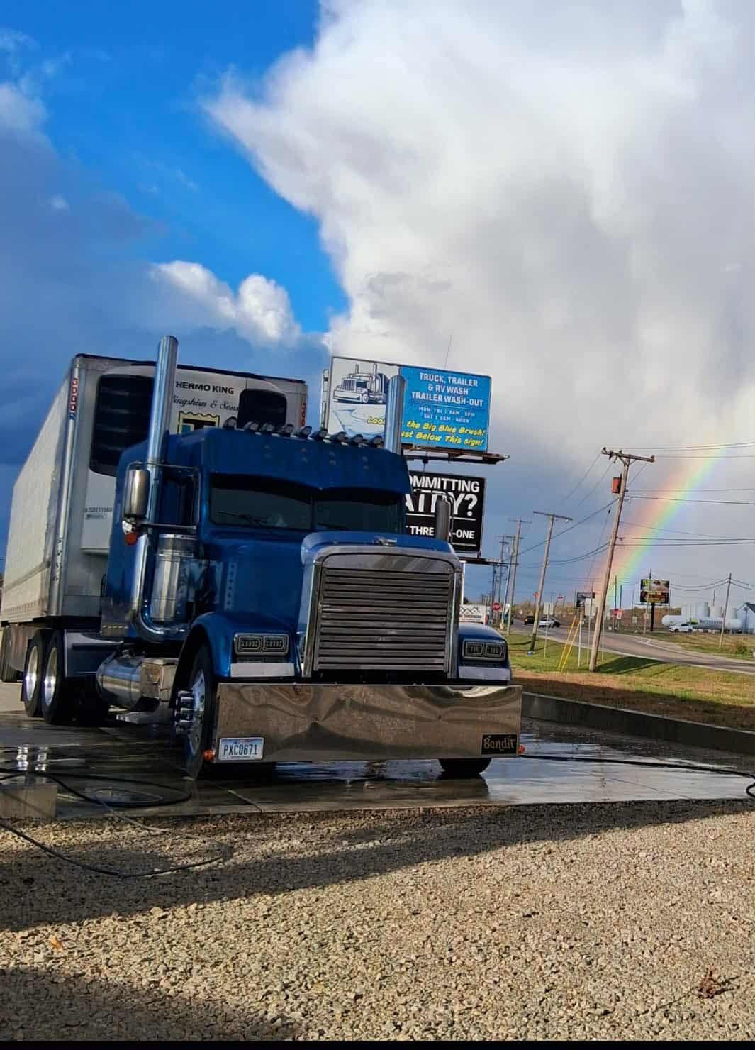 Blue semi-truck with trailer parked under a partially visible rainbow, partly cloudy sky in the background.